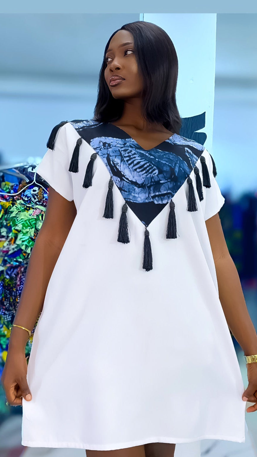 Woman wearing a white dress with black patterns and tassels in a store setting.